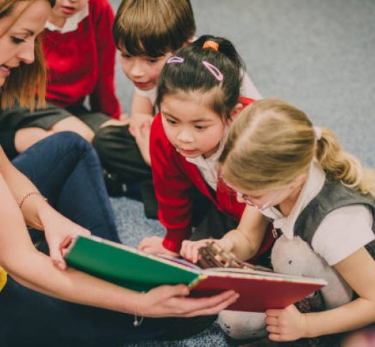 Teacher is sitting in the classroom with her primary school students, reading a story to them.