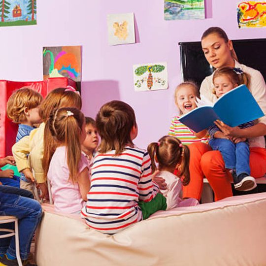 Large group of kids sit and listen to teacher reading a book and telling stories